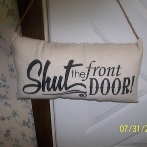 May include: Rectangular beige fabric sign with the phrase "Shut the front DOOR!" in black script. The sign is suspended by a brown rope and is hanging on a white door. The date "07/31/2013" is in the bottom right corner.