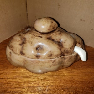 Puede incluir: Un recipiente de cerámica con forma de patata con tapa y un asa blanca en forma de colmillo. El recipiente es principalmente de color marrón claro con detalles en marrón oscuro, y la tapa tiene un pequeño pomo redondeado.