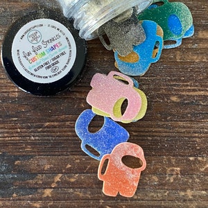 May include: A glass jar filled with silver edible glitter sprinkles, spilling out onto a wooden surface. The sprinkles are shaped like the popular video game character, Among Us. The jar label reads "fun food sprinkles custom shapes gluten free/sugar free food grade ok".