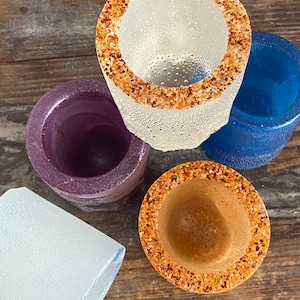 May include: Four shot glasses with different colored glass and rim finishes. One shot glass has a white glass with a brown rim, one has a purple glass with a clear rim, one has a blue glass with a clear rim, and one has a brown glass with a brown rim.