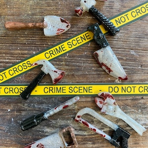 May include: A collection of seven miniature tools, including a hammer, a shovel, a knife, an axe, a screwdriver, a saw, and a wrench, all painted white with red splatter paint. The tools are arranged on a wooden surface with yellow crime scene tape.