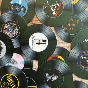 Puede incluir: Un conjunto de posavasos de vinilo negro con diseños de portadas de álbumes variados. Incluyen artistas como Nirvana, Pearl Jam y Fleetwood Mac. Los posavasos están dispuestos sobre una superficie de madera clara.