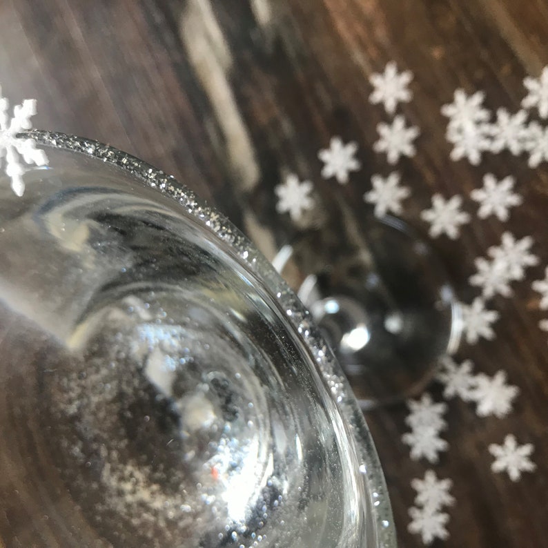 May include: A clear glass with a silver glitter rim, surrounded by white snowflake decorations on a wooden surface.