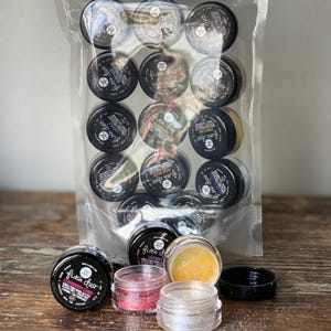 以下が含まれることがあります： 透明な袋に入った、小さな黒い蓋付きの「Flash Dust」グリッターの瓶。袋の外には、ピンク、シルバー、ゴールドなど、さまざまな色の瓶がいくつかあります。瓶のラベルには、さまざまなフレーバーや色を示すテキストが表示されています。