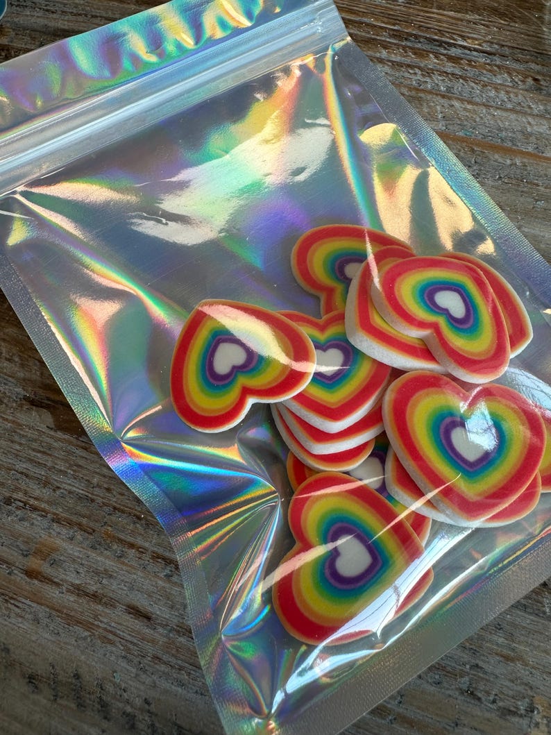 May include: A clear plastic bag filled with rainbow heart-shaped decorations. The hearts are made of a white material with a rainbow pattern.