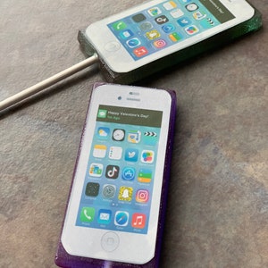 Puede incluir: Dos piruletas con forma de teléfono, de color morado y verde, con una pantalla blanca que muestra varios iconos de aplicaciones y el texto "Happy Valentine's Day!" en la pantalla.