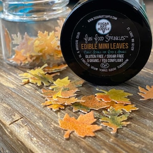 May include: A small jar of edible mini leaves sprinkles for food and drinks. The jar is black with white text that reads "fun food sprinkles" and "edible mini leaves". The jar is open and the sprinkles are scattered on a wooden surface. The sprinkles are shaped like leaves and are in shades of green, yellow, and orange.
