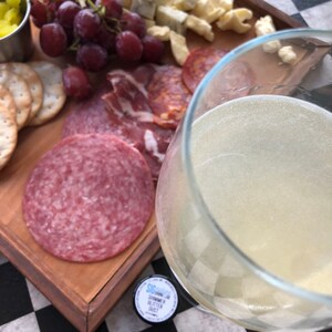 May include: A charcuterie board with salami, crackers, cheese, and grapes. A glass of shimmering liquid is in the foreground. A small container of shimmer glitter dust is visible. The board is on a black and white checkered surface.