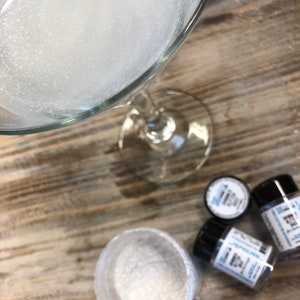 May include: Overhead shot of a clear stemmed glass filled with white foam, next to small jars of glitter and a small bowl of glitter. The jars have black lids and white labels with blue text. The items are on a wooden surface.