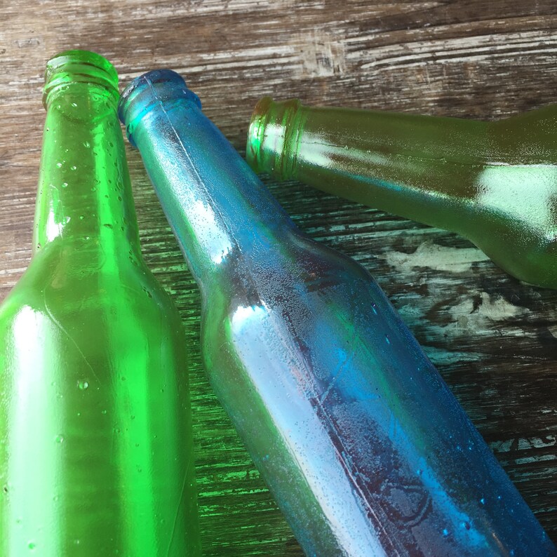 Op de afbeelding: Drie glazen flessen, &eacute;&eacute;n groene, &eacute;&eacute;n blauwe en &eacute;&eacute;n groene, liggen op een houten oppervlak. De flessen zijn allemaal van verschillende vormen en maten.