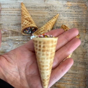 May include: Close-up of several waffle cones, some filled with chocolate and colorful sprinkles. The cones are light brown and have a textured, grid-like pattern. The image is taken on a wooden surface.