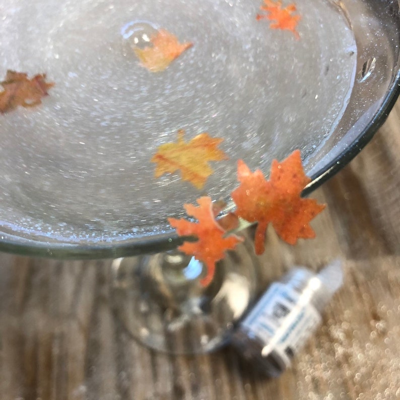 May include: A clear glass cocktail glass filled with a bubbly, light-colored liquid. Several orange and yellow leaf-shaped decorations float within the drink. The glass sits on a textured, light brown surface.