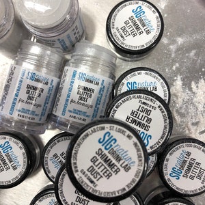 May include: An assortment of small, clear jars filled with silver shimmer glitter dust for beverages. The jars have black lids with white labels that read "Shimmer Glitter Dust" and "Signature Drink Lab". The jars are 3g (0.11 fl oz).