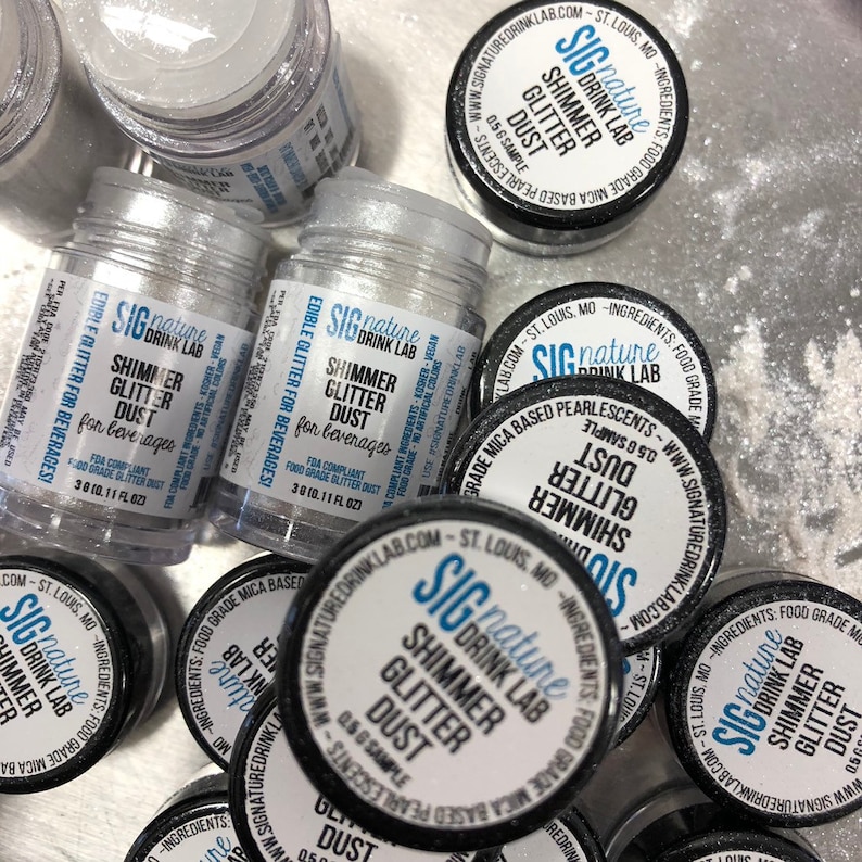 May include: An assortment of small, clear jars filled with silver shimmer glitter dust for beverages. The jars have black lids with white labels that read "Shimmer Glitter Dust" and "Signature Drink Lab". The jars are 3g (0.11 fl oz).