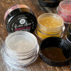 以下が含まれることがあります： 「Strawberry Candy」と書かれた「Flash Dust」の文字が入った、食用グリッターの瓶がいくつかあります。グリッターの色は様々で、ピンク、ゴールド、クリアなものがあります。瓶は透明で、黒い蓋が付いています。