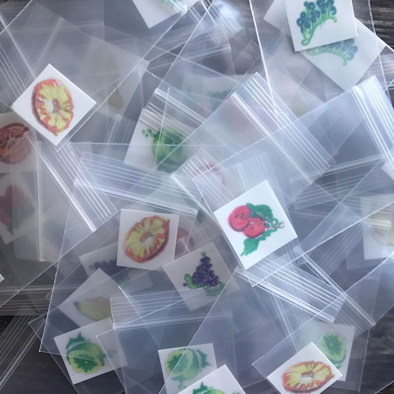 Pode incluir: Uma pilha de sacos de pl&aacute;stico transparentes contendo pequenos adesivos coloridos com imagens de frutas, vegetais e outros alimentos.