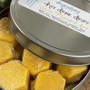 May include: A silver tin with a lid containing six hexagonal golden margarita lime drink drops. The lid has a label that reads "Signature Drink Lab Hex Drops Golden Margarita Lime Add to sparkling water for instant glittering drinks!"