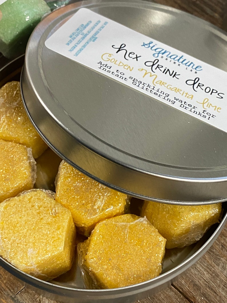 May include: A silver tin with a lid containing six hexagonal golden margarita lime drink drops. The lid has a label that reads "Signature Drink Lab Hex Drops Golden Margarita Lime Add to sparkling water for instant glittering drinks!"