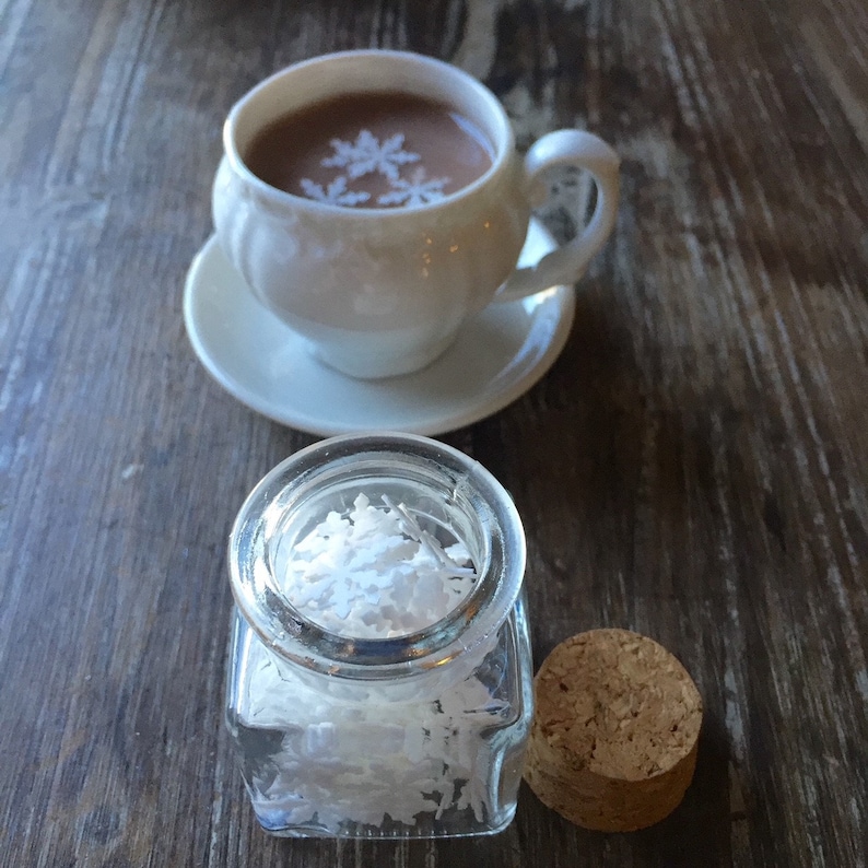 May include: A white ceramic mug with a brown liquid inside. The liquid has white snowflake designs on top. The mug is sitting on a white saucer. A small glass jar with a cork lid is filled with white shredded coconut.