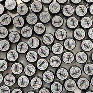 May include: Overhead shot of numerous small, black containers with white lids. Each lid features a colorful label with text, including "SHIMMER GLITTER DUST" and color names like "BLUE" and "RED". The containers are arranged closely together.