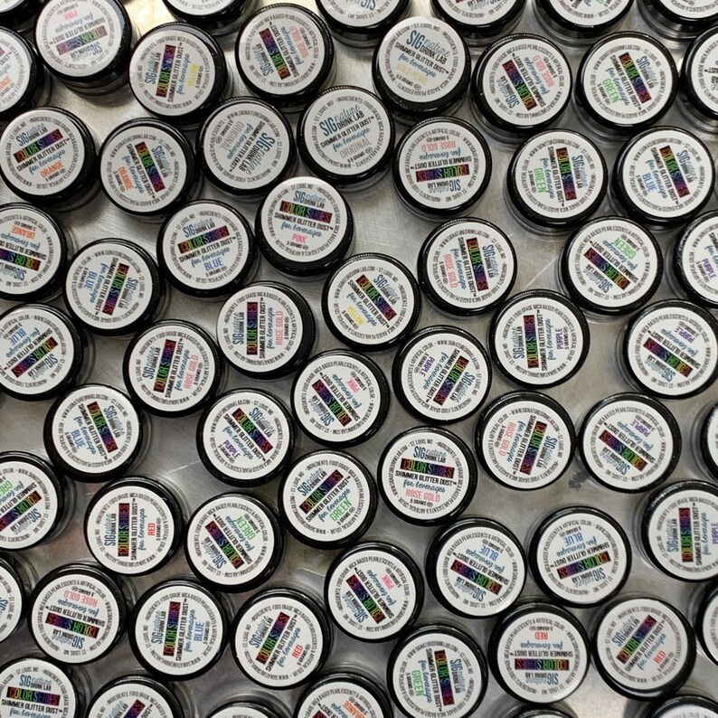 May include: Overhead shot of numerous small, black containers with white lids. Each lid features a colorful label with text, including "SHIMMER GLITTER DUST" and color names like "BLUE" and "RED". The containers are arranged closely together.