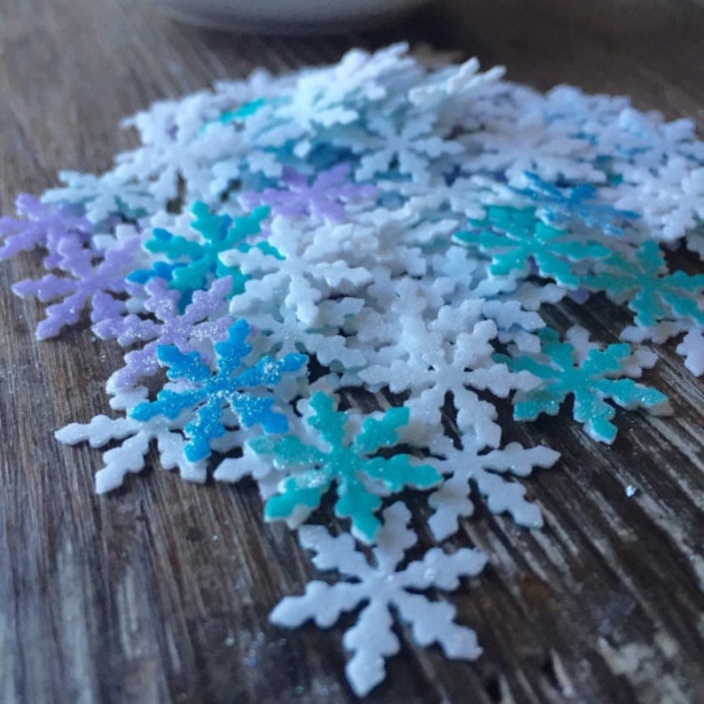 May include: A pile of white, blue, and purple glitter snowflake sprinkles. The snowflakes are scattered on a wooden surface.