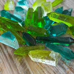 Puede incluir: Una colección de cristales de vidrio facetados y coloridos en tonos verdes y azules. Los cristales varían en tamaño y forma, algunos parecen translúcidos y otros más opacos. Los cristales están dispersos sobre una superficie de madera.