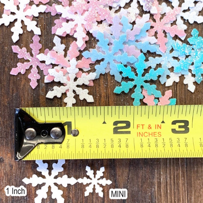 May include: A close-up of a measuring tape showing 1 inch and 2 feet.  The tape is measuring a variety of small, glittery snowflake shapes in pink, blue, and white.  The snowflakes are labeled "MINI".