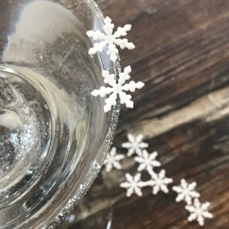 May include: A glass with a silver rim and white snowflake sprinkles around the edge. The glass is filled with a clear liquid. There are more white snowflake sprinkles on a wooden surface.