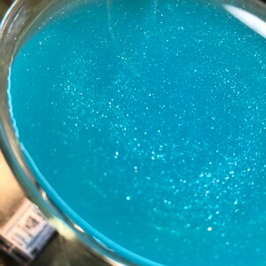May include: A close-up view of a cocktail in a clear glass. The drink is a vibrant, sparkling turquoise color, with tiny shimmering particles throughout. The glass is partially visible, showing the liquid's edge and the rim.
