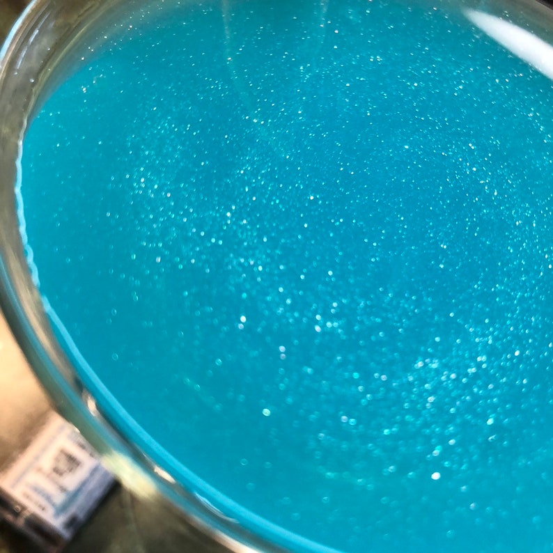 May include: A close-up view of a cocktail in a clear glass. The drink is a vibrant, sparkling turquoise color, with tiny shimmering particles throughout. The glass is partially visible, showing the liquid's edge and the rim.
