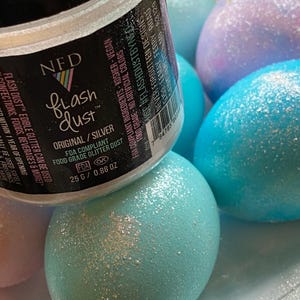 以下が含まれることがあります： 「Flash Dust」とラベルされた銀色の食用グリッターの瓶が、パステルカラーのグリッターをまぶした卵の上に置かれています。卵は青、ピンク、紫の色合いです。瓶のラベルには、食品グレードでビーガンであることが記載されています。