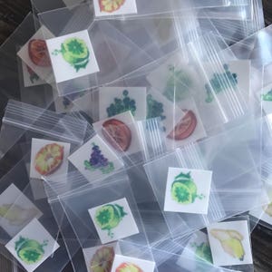 Pode incluir: Uma variedade de adesivos coloridos de frutas e vegetais em pequenos sacos de pl&aacute;stico transparentes. Os adesivos apresentam imagens de lim&otilde;es, limas, laranjas, uvas e outras frutas.