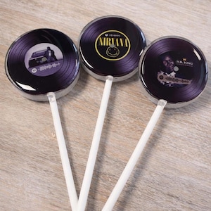 May include: Three round lollipops with a vinyl record design. Each lollipop features a different image: a man with a car, the Nirvana logo, and B.B. King. The lollipops have white sticks and a glossy finish.