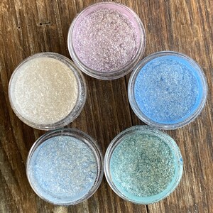 以下が含まれることがあります： キラキラと輝くカラフルなパウダーが入った、小さな透明なプラスチック製の瓶が5つ。色は、ライトピンク、ライトブルー、白、ティールグリーンなど。木製の表面に円形に配置されています。