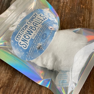 May include: A clear plastic bag with a white cotton candy snowball inside. The bag has a label that says "Signature Cotton Candy Snowballs for Drinks". The label also has a FDA OK symbol and a website address.