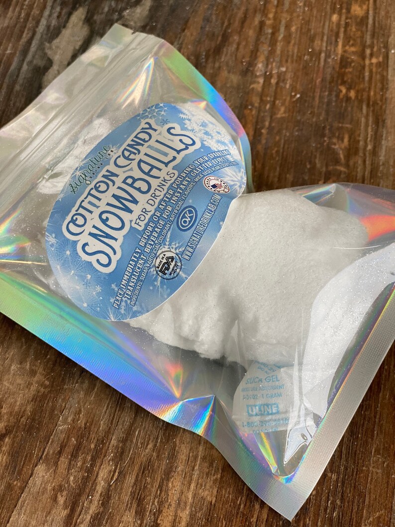 May include: A clear plastic bag with a white cotton candy snowball inside. The bag has a label that says "Signature Cotton Candy Snowballs for Drinks". The label also has a FDA OK symbol and a website address.