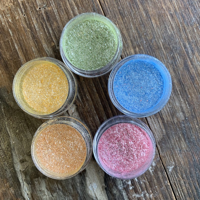 以下が含まれることがあります： カラフルなグリッターが入った、小さな透明なプラスチック製の瓶が5つ。 グリッターの色は、ゴールド、グリーン、ブルー、ピンク、オレンジです。 瓶は、風化した木製の表面に円形に配置されています。
