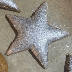 以下が含まれることがあります： 銀色のグリッターで覆われた星型のクッキーのクローズアップ。クッキーは薄茶色の表面にあり、背景には他の星型のクッキーが見えます。クッキーは五芒星です。