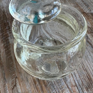 May include: A clear glass jar with a lid. The jar has a rounded bottom and a wide mouth. The lid has a knob on top.