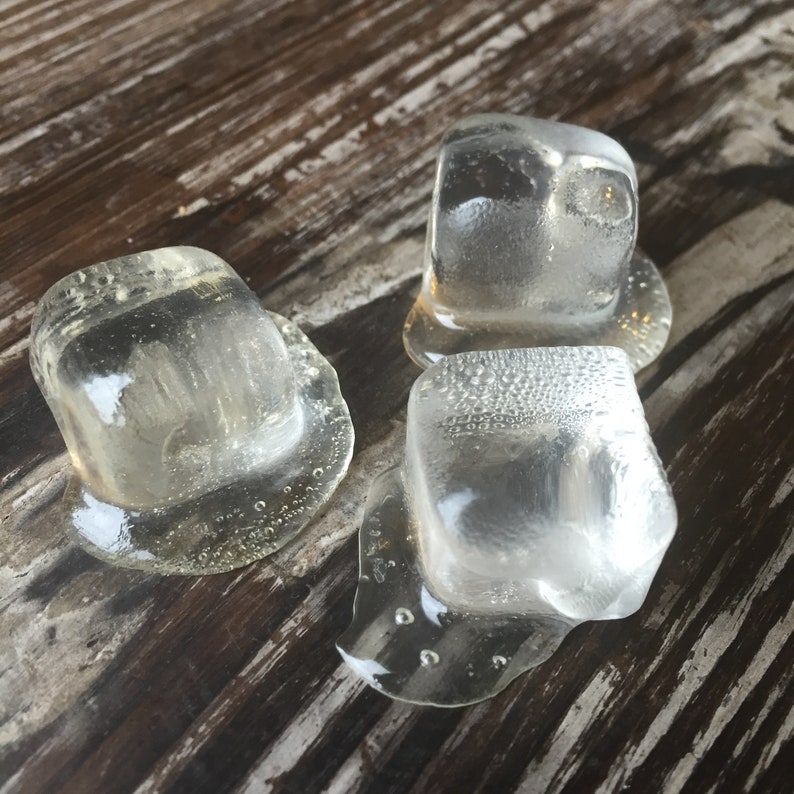 Op de afbeelding: Drie heldere ijsblokjes smelten op een houten oppervlak. De ijsblokjes zijn licht bevroren en hebben een doorschijnende uitstraling.