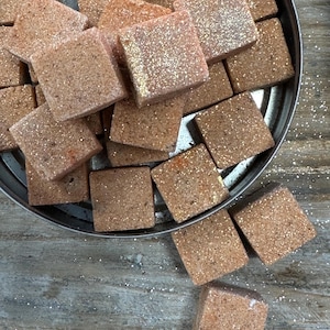 May include: A collection of square, brown bath bombs with glitter on a silver tray. The bath bombs are arranged in a pile, with some spilling out onto a wooden surface. The glitter adds a touch of sparkle to the otherwise earthy tones.
