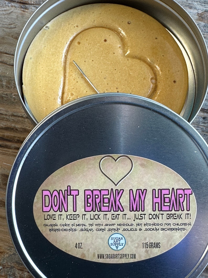 May include: A black metal tin with a label that reads 'Don't Break My Heart' in pink letters. The label also includes the text 'Love it, keep it, lick it, eat it... just don't break it!' in smaller pink letters. The label also includes the text 'Dalgona candy in metal tin with sharp needle. Not intended for children. Ingredients: sugar, corn syrup solids & sodium bicarbonate.' in smaller black letters. The label also includes the text '4 oz.' and '115 grams' in black letters. The label also includes the text 'Sugar Art Supply' in white letters on a pink background. The label also includes the text 'www.sugarartssupply.com' in black letters. The tin contains a brown, heart-shaped candy.