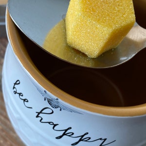 May include: A white ceramic mug with a gold rim and the text "bee happy" in black script. A silver spoon holds a yellow honeycomb-shaped sugar cube.