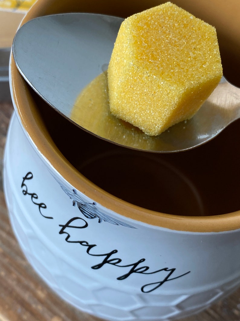 May include: A white ceramic mug with a gold rim and the text "bee happy" in black script. A silver spoon holds a yellow honeycomb-shaped sugar cube.