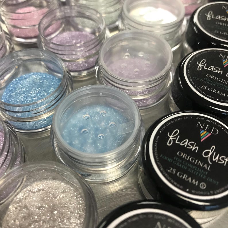 以下が含まれることがあります： カラフルなグリッターが入った、小さな透明なプラスチック製の瓶の詰め合わせ。瓶には、青、ピンク、紫、銀色のグリッターが入っています。一部の瓶は開いており、その他は「Flash Dust」と「25 Gram」と書かれた黒い蓋で閉じられています。