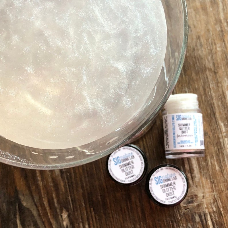 May include: A clear glass bowl filled with a shimmering, pearlescent liquid. Three small jars of shimmer glitter dust are next to the bowl. The jars have labels that read "SIG nature's DRINK LAB SHIMMER GLITTER DUST".