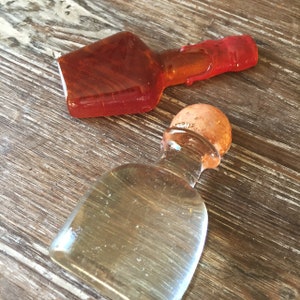 Op de afbeelding: Twee glazen flessen, een amberkleurig en een helder, met kurken stoppen. De amberkleurige fles heeft een rode dop. De flessen staan op een houten oppervlak.