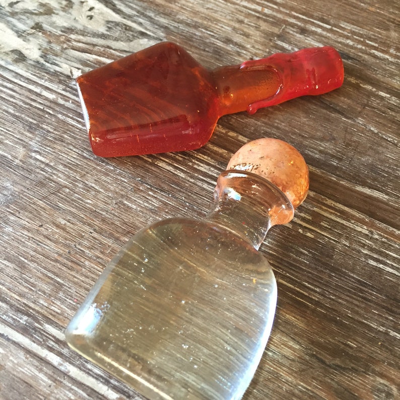 Op de afbeelding: Twee glazen flessen, een amberkleurig en een helder, met kurken stoppen. De amberkleurige fles heeft een rode dop. De flessen staan op een houten oppervlak.