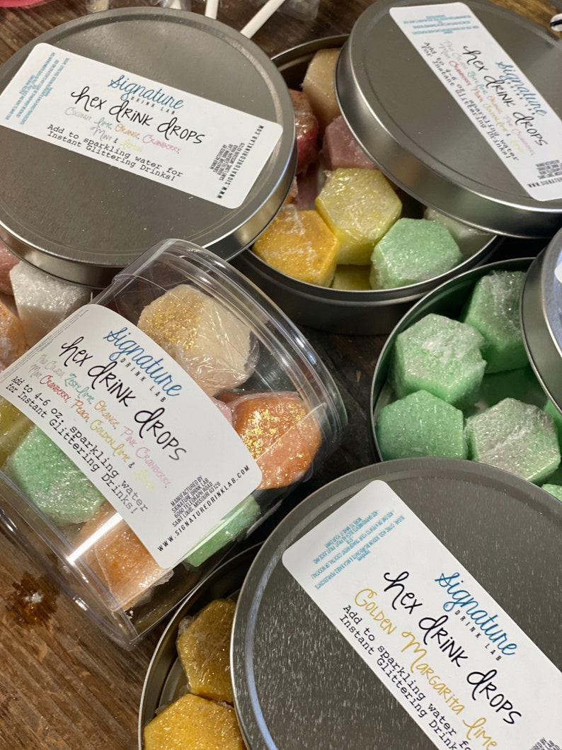 May include: Three silver tins filled with colourful hexagonal drink drops. The tins have labels that read "Signature Drink Lab Hex Drink Drops" with different flavour combinations: "Coconut Peach, Golden Lime, Mint & Orange".